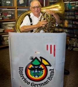 Harry Wolf of the Waterloo German Band is seen here at a music stand with his century-old Czechoslovakian tuba. The cataloged plastic boxes behind him contain more than 40 pairs of lederhosen that he wears for performances. (Alan Dooley photo)